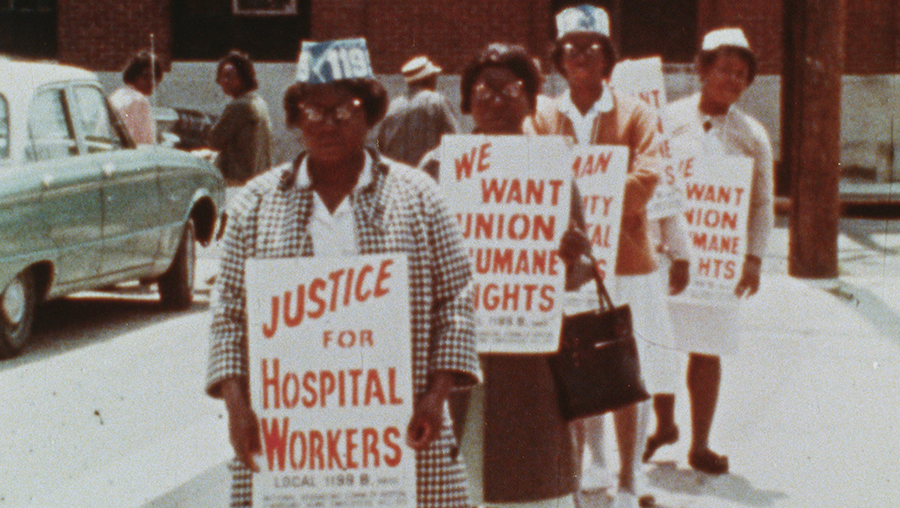 A group of Black women, wearing hats indicating that they are Local 1199 B union members, march in front of their workplace carrying signs reading "Justice for Hospital Workers" and "We Want Union Humane Rights"